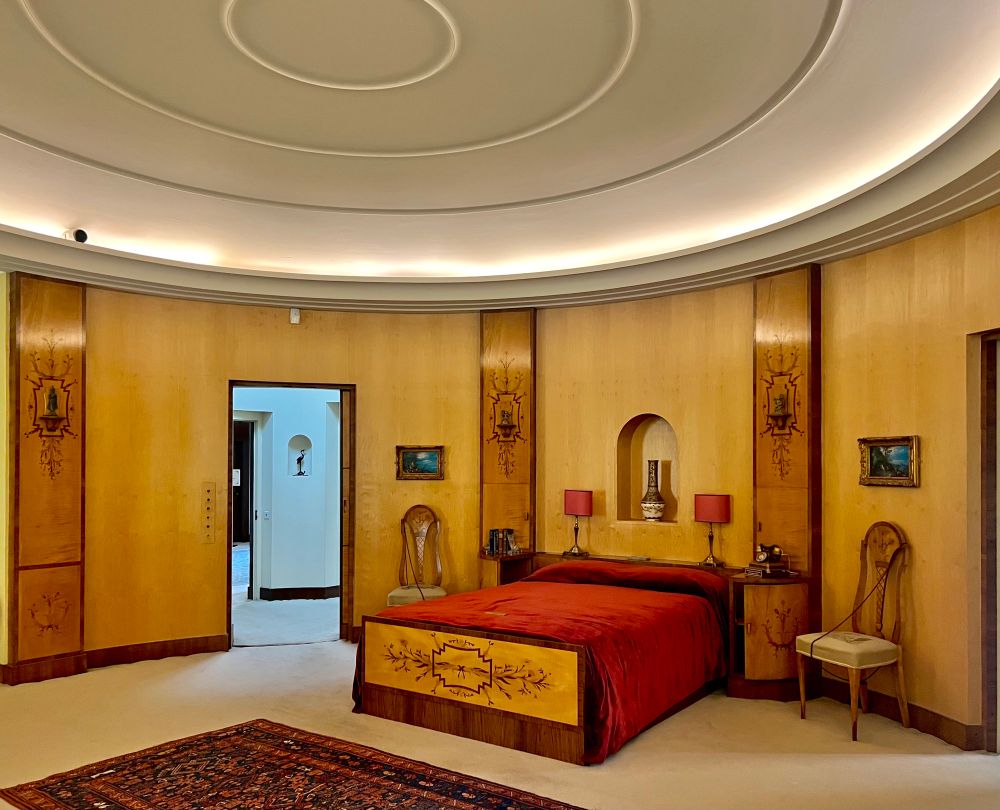 A bed with red bedding underneath a ribbed circular ceiling. The walls are wood and there is an open doorway leading to a hallway.