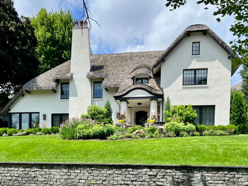 Large white Storybook style house sitting atop a grassy hill with a stone retaining wall at the bottom.