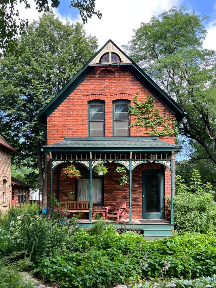 A simple red brick two-story, front gabled house with a decorative porch featuring carved woodwork, surrounded by lush greenery.