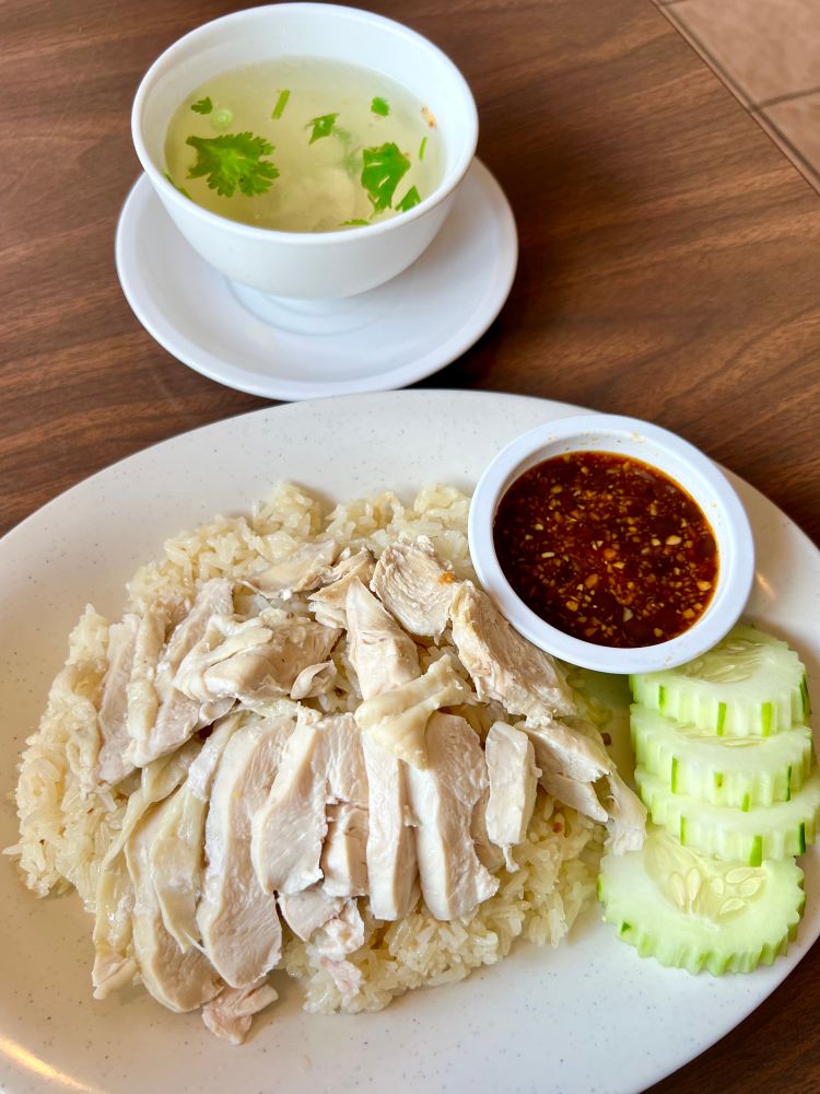A plate of rice topped with slices of chicken. The plate also has a bowl of dark sauce and sliced cucumbers. There is a bowl of chicken broth with cilantro next to the plate.