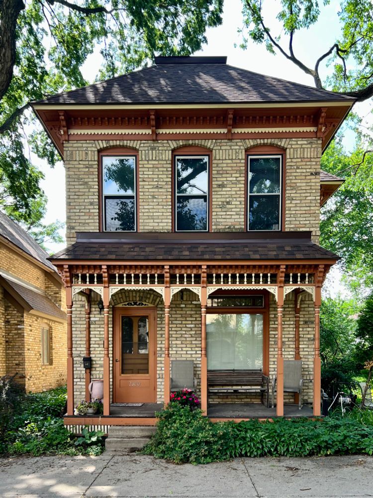 A simple two-story house with an Italianate roof and a decorative porch featuring carved woodwork, surrounded by lush greenery.