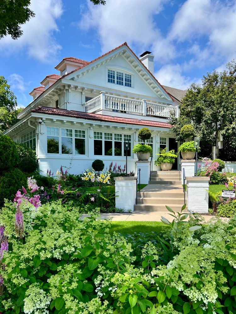 Eclectic revival house with red tile roof situated within a lush garden of flowers and greenery.
