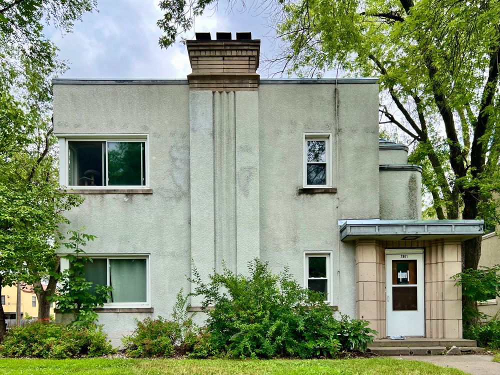 Boxy stucco duplex with a fluted chimney, recessed doorway, and stepped walls above the entrance.