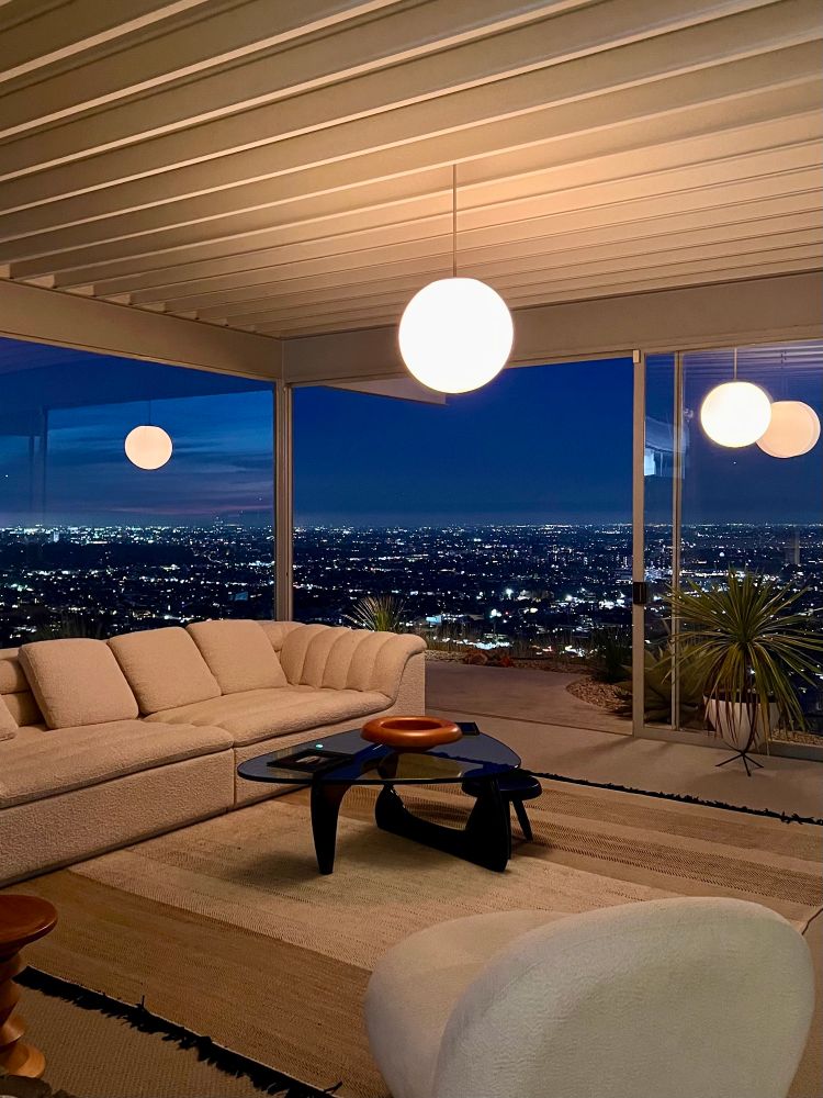 Inside the living room of the house looking out over Los Angeles at night. One of the glass walls is pulled back. There is a large globe light pendant in the center of the ceiling and a white couch to the left of the room. 