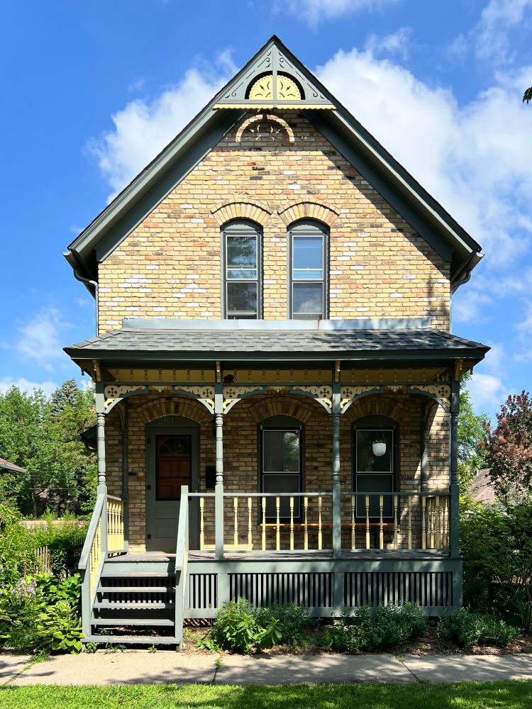 A front gable brick house with a porch surrounded by greenery.
