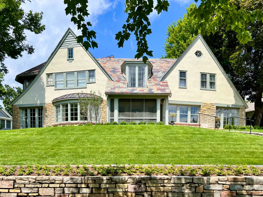 Dual gabled English style home with a multicolored slate roof sitting atop a grassy hill with a stone retaining wall below.
