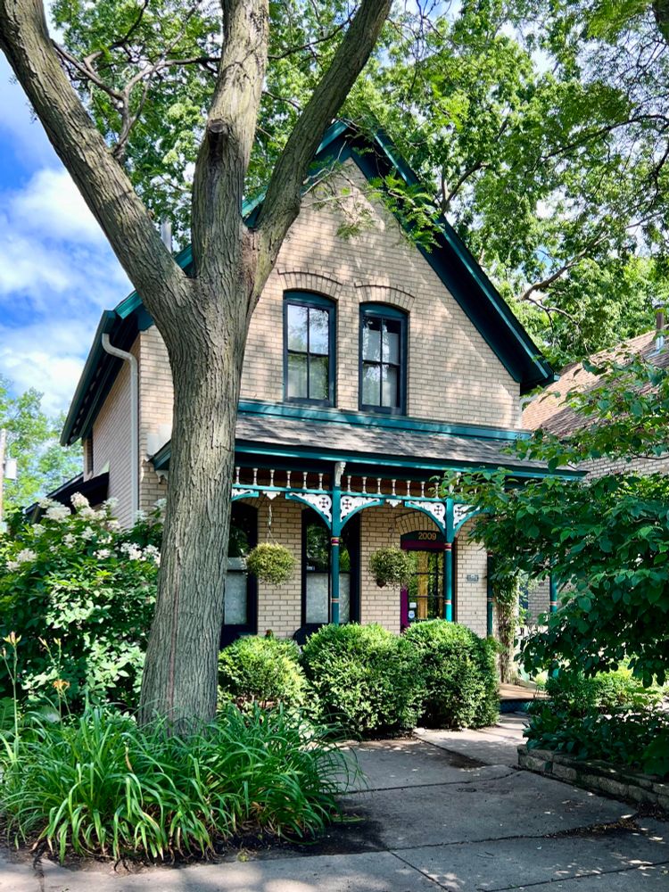 A front gable brick house with a blue, ornate porch surrounded by greenery.
