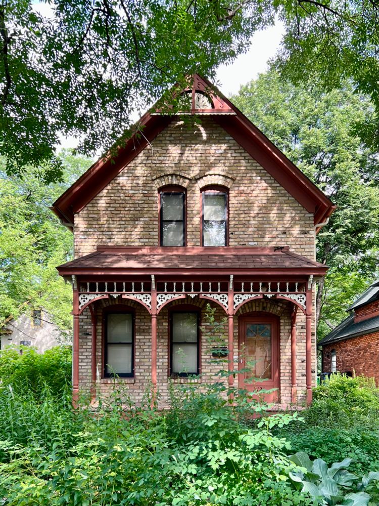 A simple two-story, front gabled house with a decorative porch featuring carved woodwork, surrounded by lush greenery.
