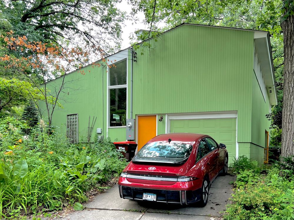 Green midcentury house with an asymmetrical gable roof. The house has an orange door and only two windows. There is a red car in the driveway.