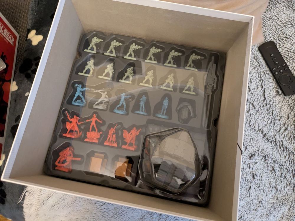 A photo of the inside of the boardgame box, it features custom molded plastic around the pieces, has several small figures of different characters, bosses, cardboard boxes and a figure of Metal Gear Rex, a huge mech.