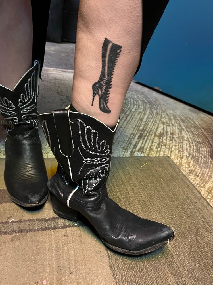 A person’s legs wearing cowboy boots, one of which is pulled down to show a tattoo on their ankle of a black leather high heeled boot based on a photo from Bizarre Magazine