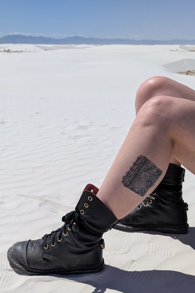 A pair of legs wearing black leather boots with a tattoo of the magic book from the fil Hocus Pocus, sitting in the sand. 