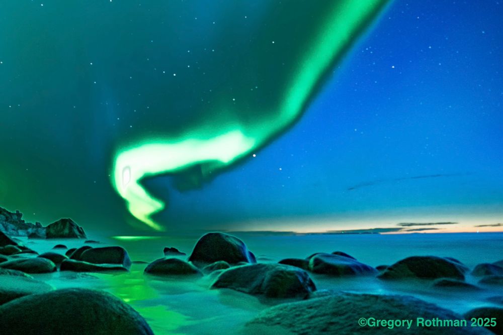 This image depicts a nighttime landscape of the Northern Lights (Aurora Borealis) over a rocky shoreline. The sky glows in vivid shades of green and turquoise as the aurora forms a sweeping, ribbon-like curve that twists gracefully across the sky. The stars are faintly visible in the deep blue background, and the horizon glows with a soft golden hue, hinting at either dawn or dusk. Smooth, rounded rocks in the foreground are bathed in the aurora’s reflection, giving the water and stones a surreal greenish glow. The overall atmosphere is serene and otherworldly. In the bottom right corner, the text “©Gregory Rothman 2025” 