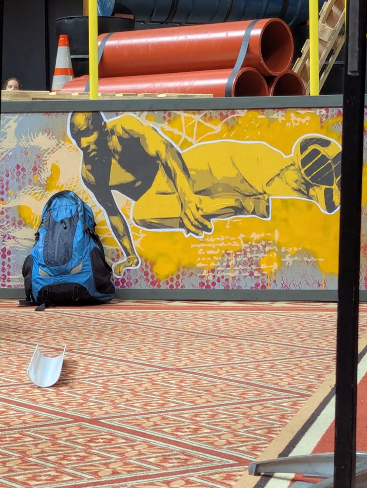 A close up of a yellow, grey, and pink side-banner along an obstacle course. The artwork depicts a male individual, Sebastien Foucan, vaulting over an obstacle.