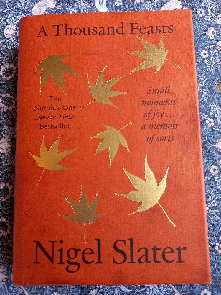 Photo of A Thousand Feasts by Nigel Slater, a burnt orange hardback with gold maple leaves. William Morris tablecloth background. I love chintz.