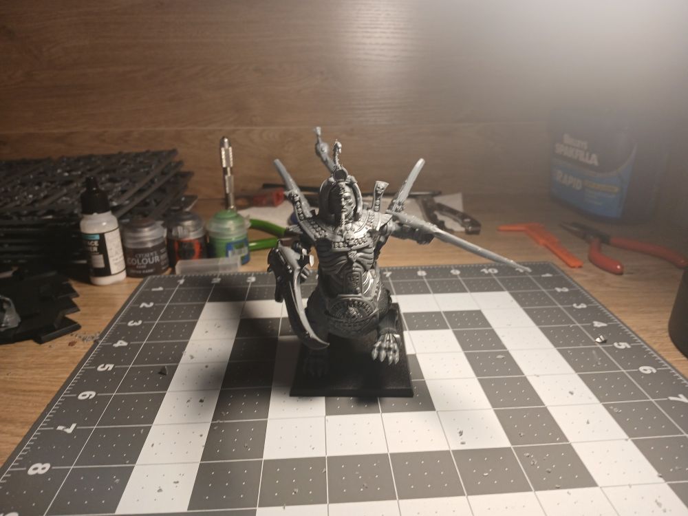 A photo showing an unpainted model depicting centaur like creature but with a lion's body. The creature has a man's face with an ancient egyptian style beard and long blades attached to the arms on the human torso. This model sits on a cluttered workbench, with tools and paint pots scattered around it in the background. 