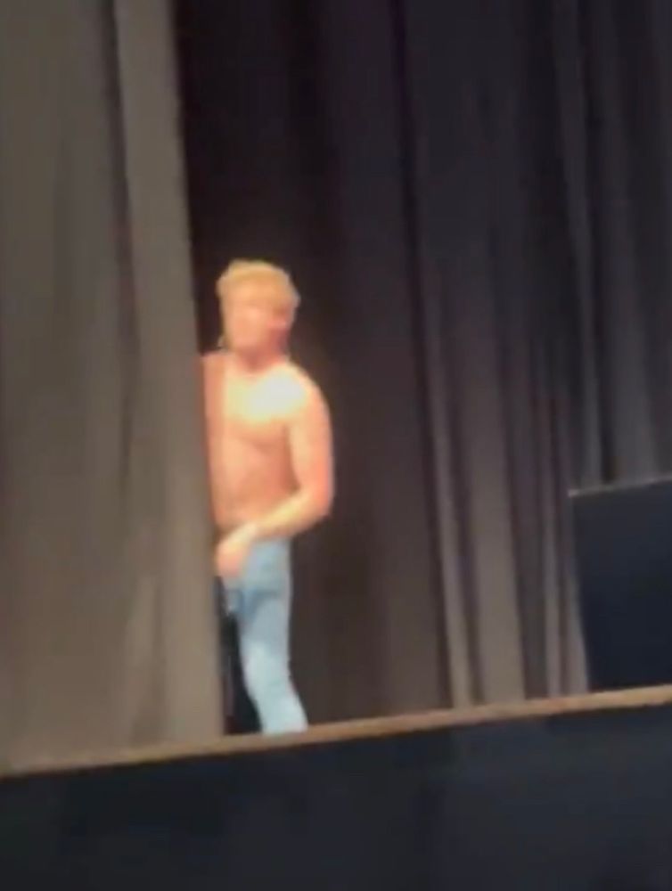 Rhys Darby on stage, topless, sneaking a look back to the audience behind the curtain