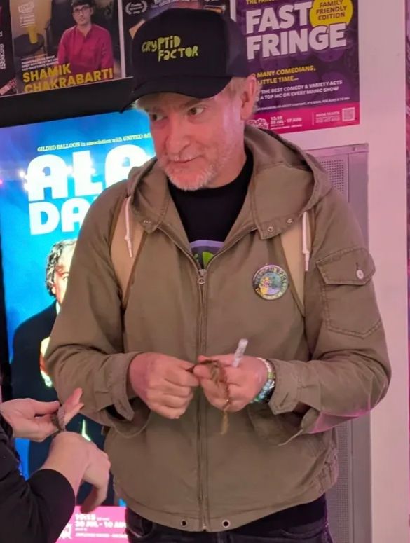 Rhys Darby in a black cap and green jacket holding some bracelets 