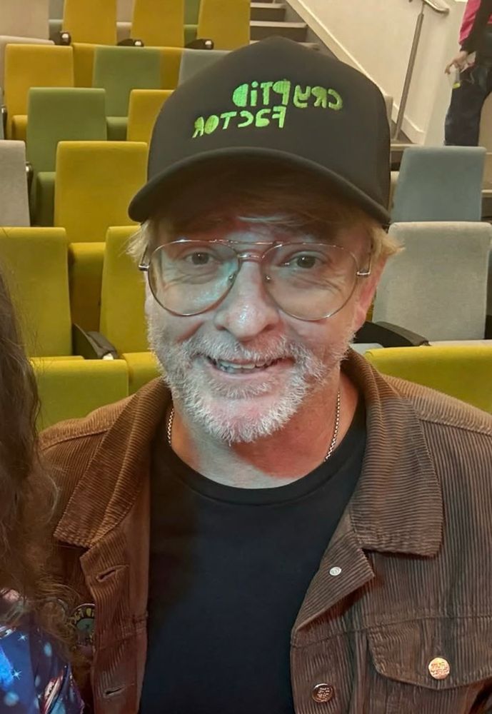 Rhys Darby wearing a cap, glasses and a black top with a brown jacket 