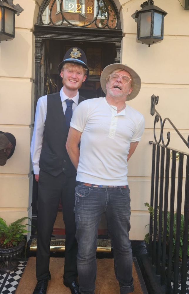 Rhys Darby in a white top and black jeans being “arrested” outside of 221B Baker Street