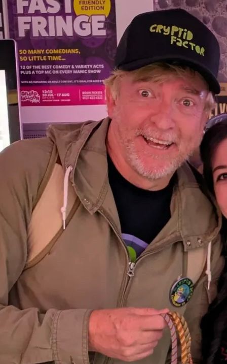 Rhys Darby in a black cap and green jacket holding some bracelets 