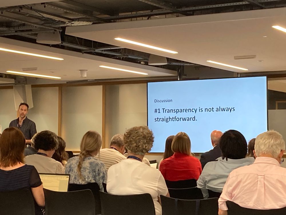 Tom standing at a podium with a slide behind him saying “Transparency is not always straightforward”