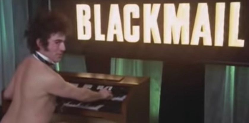 Monty Python's Terry Jones, naked, at an organ. A large, lit sign behind him reads "BLACKMAIL"