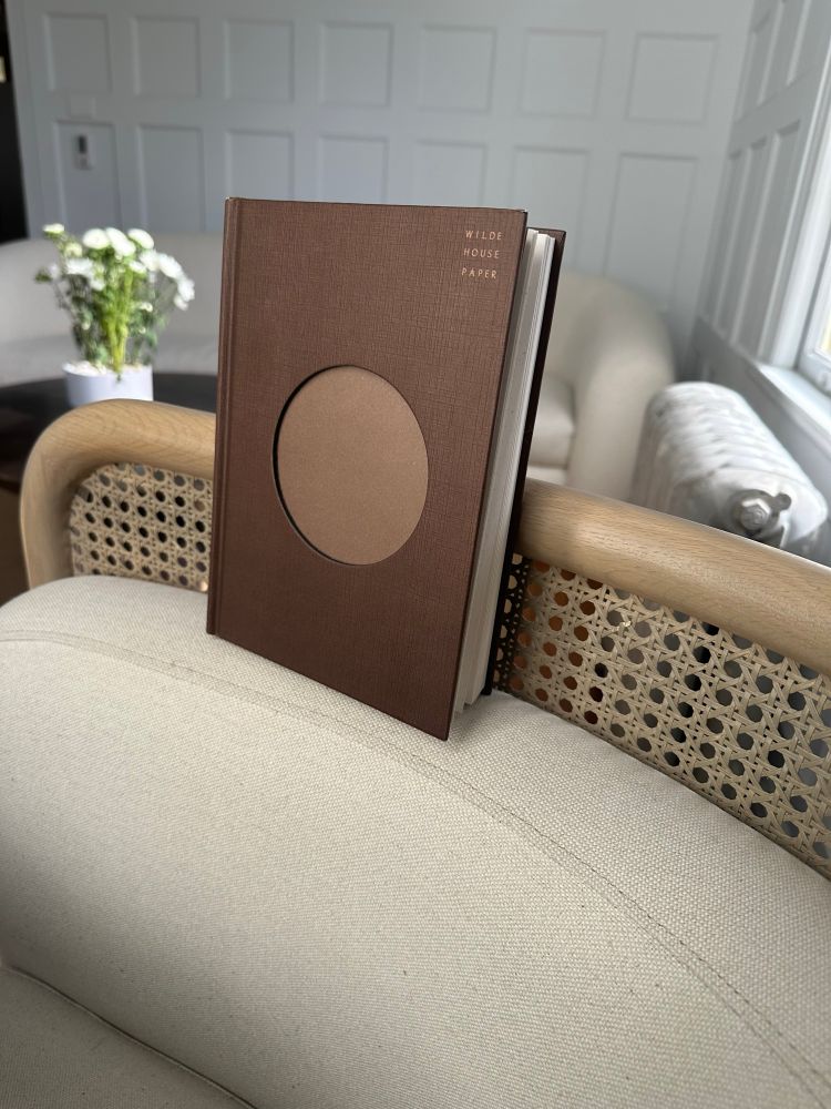 Image of a brown journal with a circular cut out in the middle propped on a beige and white chair in a room with a cream colored couch and white flowers in the background