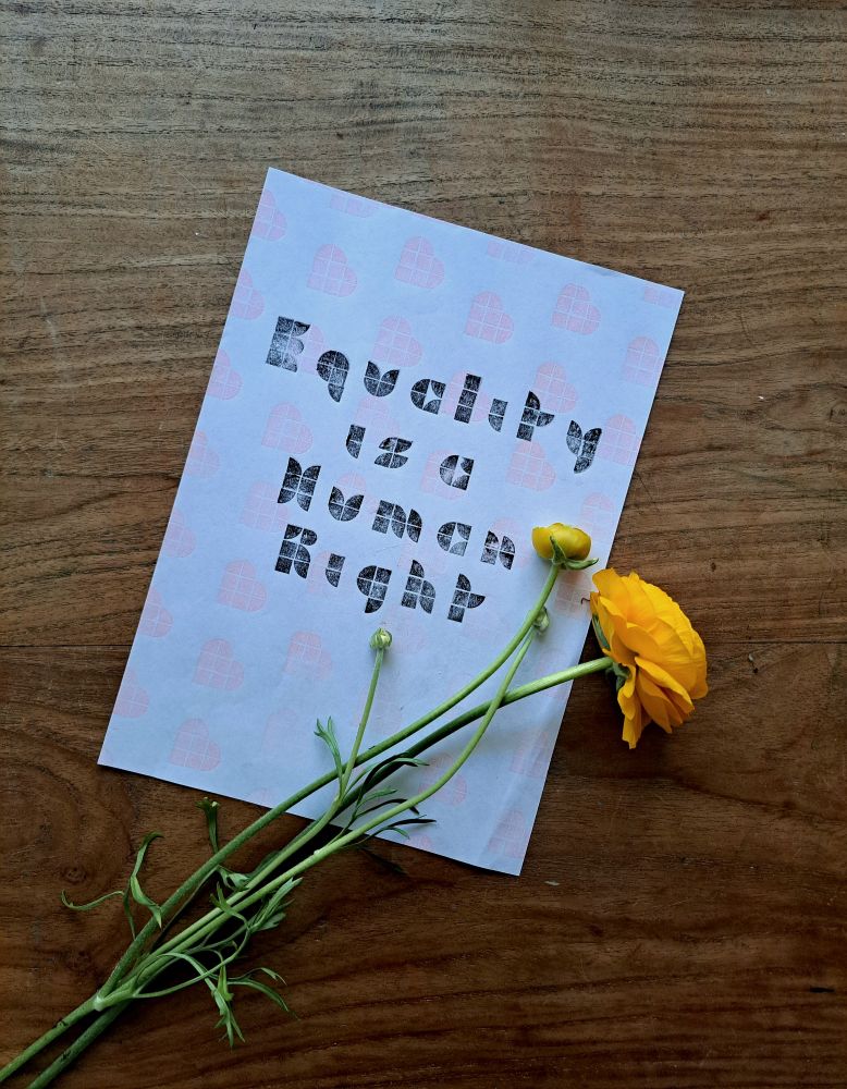 A4 Print made with dots lego. It says 'equality is a human right' in black. On the background of the print there are pink hearts, also printed with dots lego. The print lies on a wooden table, and yellow flower lies on right corner of the print.
