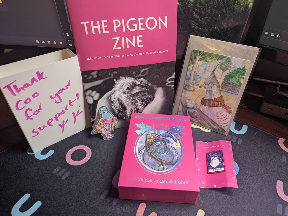 A pink box with holographic accents, with a pigeon in a wizard hat on the center. It's labeled "The Pigeon Oracle: Guidance from a Dove". Nearby are a pin and sticker with similar pigeons, a piece of art depicting a crested pigeon in a scarf enjoying coffee and a croissant, and a booklet labeled 'The Pigeon Zine'. A piece of cardboard packaging material reads "Thank coo for your support!"