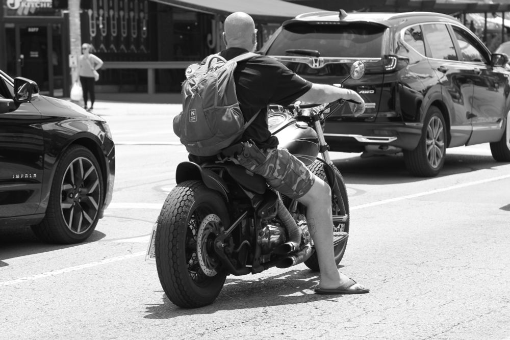 A black and white photo of a man on a Harley Davidson motorcycle with open pipes. He is wearing shorts, a tee shirt, flip flops, and no helmet. He is openly carrying a stainless revolver in an outside-the-waistband holster.