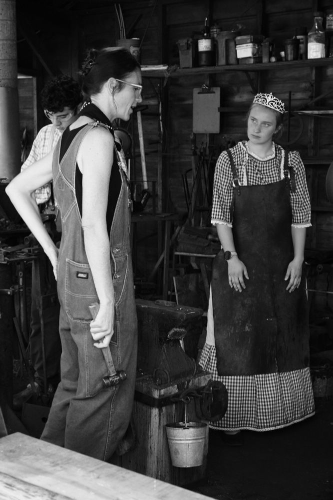 A young woman in a checked dress, tiara, and leather apron listens intently as a woman in overalls and holding a hammer stands at an anvil and explains blacksmithing