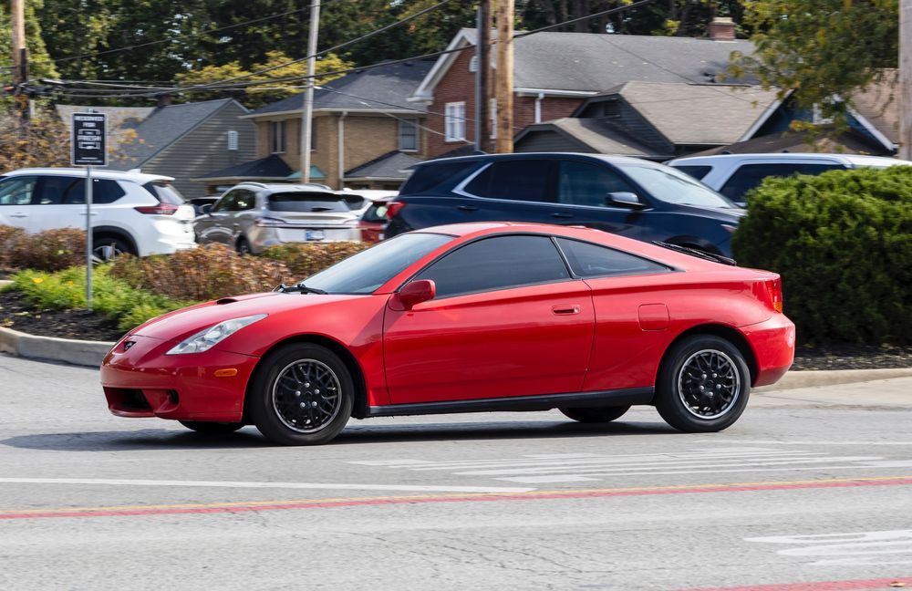 A red '00-'02 Toyota Celica driving down a city street.