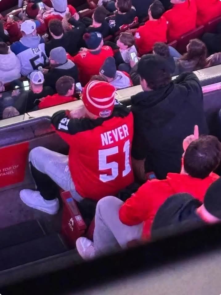 A hockey game where fans are sitting in the stands and we see them from the back. One guy is wearing a red jersey that says Never 51.