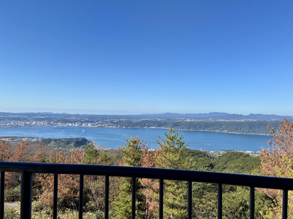 展望台の手すり越しに見える青い海と湾岸の街並み、奥には山々が連なり、快晴の空が広がる風景。