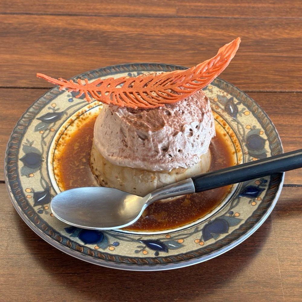 おしゃれな皿にのったチョコプリン、チョコ風味生クリームがたっぷりのって、1番上にはプリンからはみ出す大きさの朱色で羽根の形をした繊細なクッキーがのっている