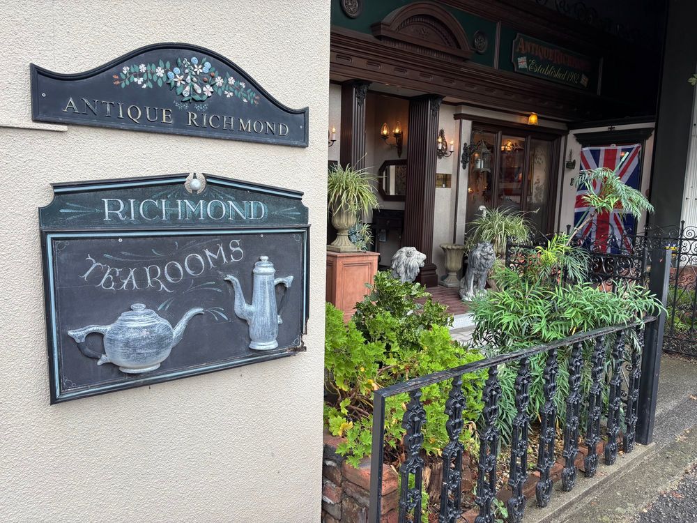 喫茶店の入り口を左斜めから
手前の壁には看板が掲げられており、店名とtea roomの文字と立体的なティーポットのイラスト
鉄の柵と植物、イギリス国旗が見える