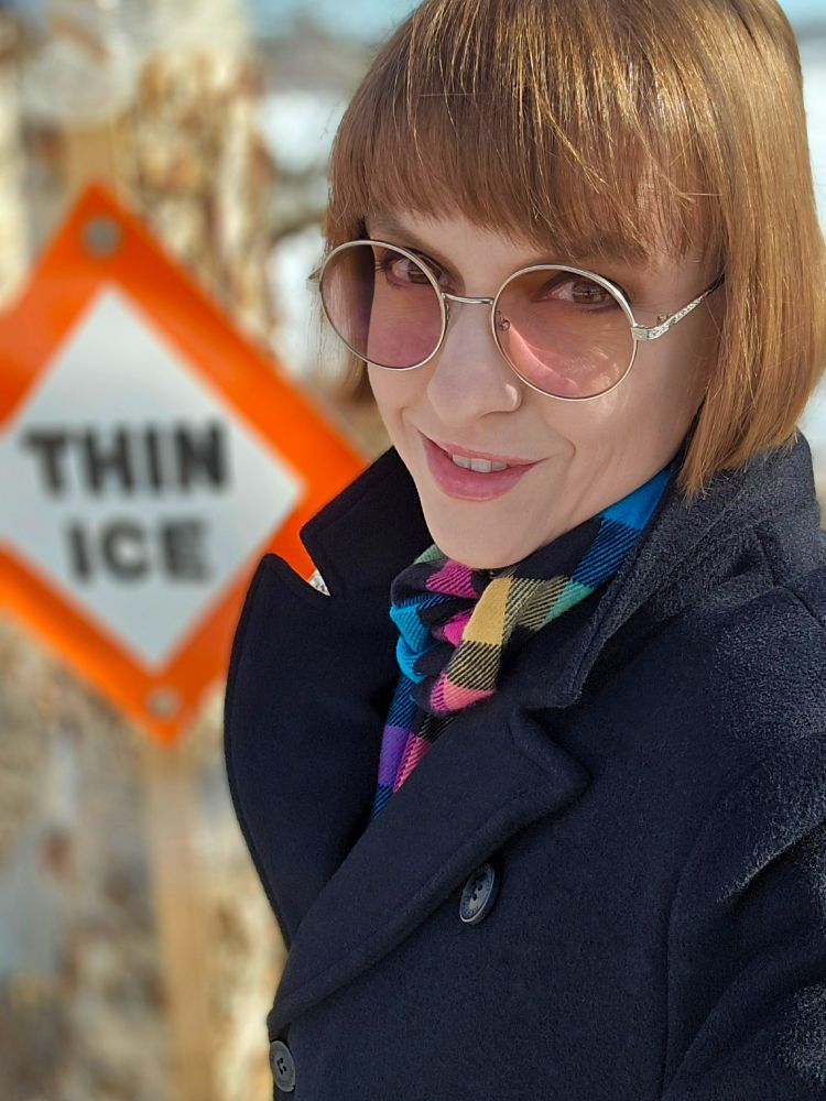 Photo of a gremlin wearing a black overcoat, with CMYK-plaid scarf and rose-tinted glasses, in front of a "Thin Ice" sign.