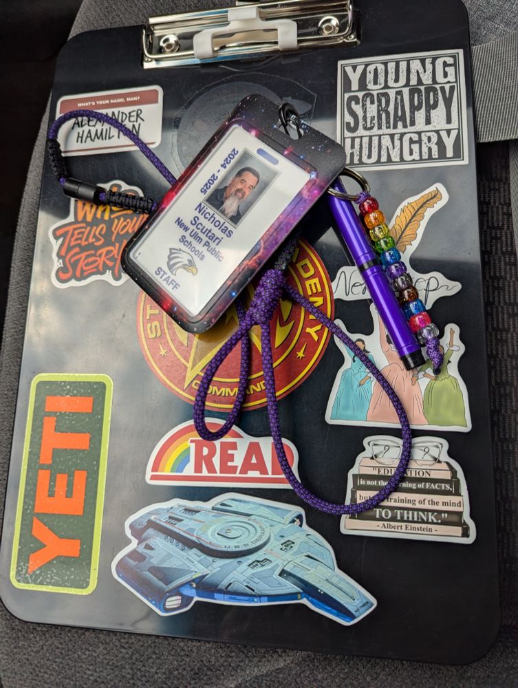 A photo of Nick's clipboard he uses for school, with his ID lanyard on top. A Rainbow bead keychain, and purple Space Pen can also be seen.