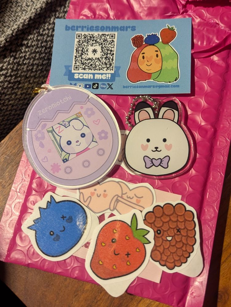 A set of charms, one shaped like a tamagotchi in pastel colors with a rabbit in the 'screen'. Another charm is a white bunny head with a little lilac bow. Includes some cute fruit stickers (a blueberry, strawberry and raspberry) and a little sticker of a pair of hearts, one hugging the other. Created by @berriesonmars.bsky.social