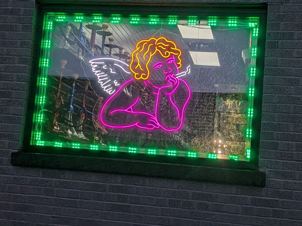 An amazing neon rendition of a cherub posed as if it's leaning on a table... smoking in the window of a smoke shop. Magnificent. 