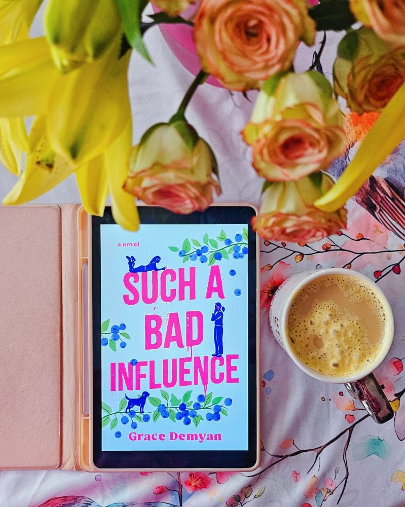 Samsung tablet in a pink case open to the cover of Such a Bad Influence by Grace Demyan. A cup of coffee with melted whipped cream is next to it, and dual colored roses and yellow lilies are closer to the camera above the ebook and mug.  