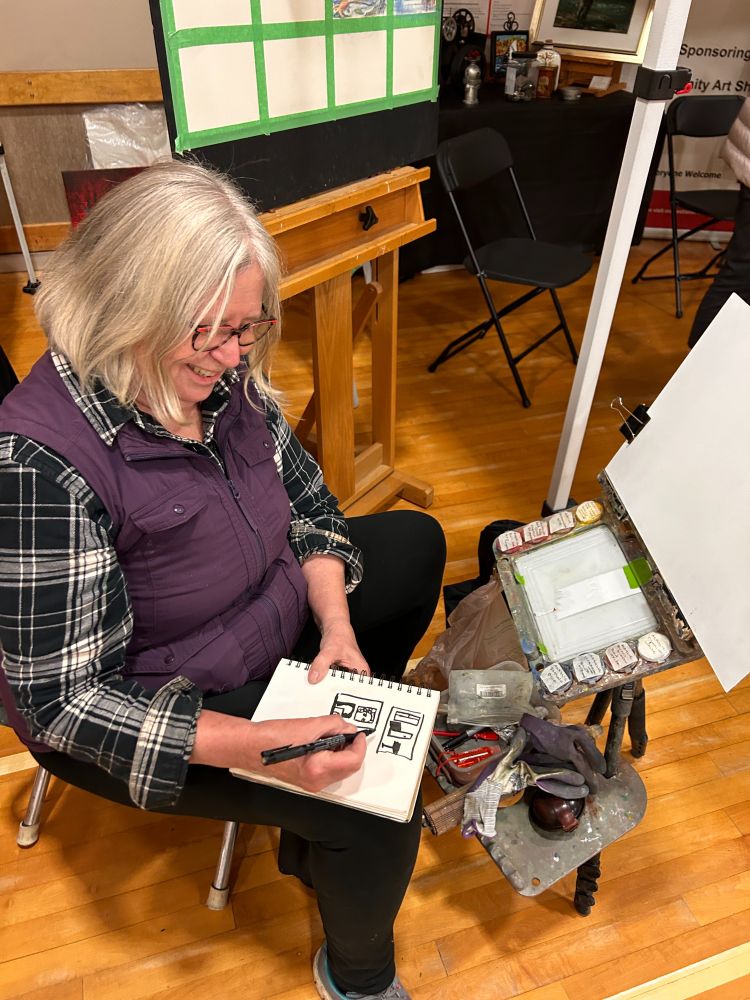 An artist sketching with marker on a sketchpad. She is sitting on a small stool with a plein air artist easel and palette set up. Photo of Artist Pat Palmer. 