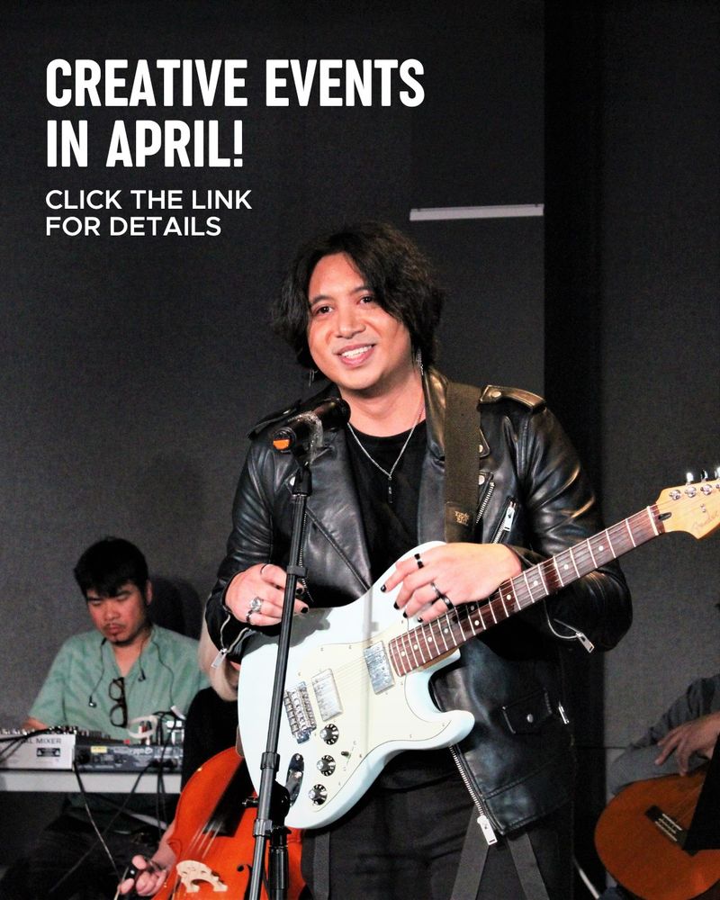 A performer holding a guitar while smiling standing at a microphone. People playing instruments in the back. Photo by Herbert Fong.