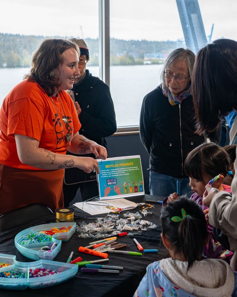 A facilitator wearing an orange "Every Child Matters" t-shirt showing a group of kids and parents how to do an activity. Photo credit: Family Fun Zone at RiverFest 2024 organized by The Fraser River Discovery Centre Society. Photo by Jonathan Garsson.