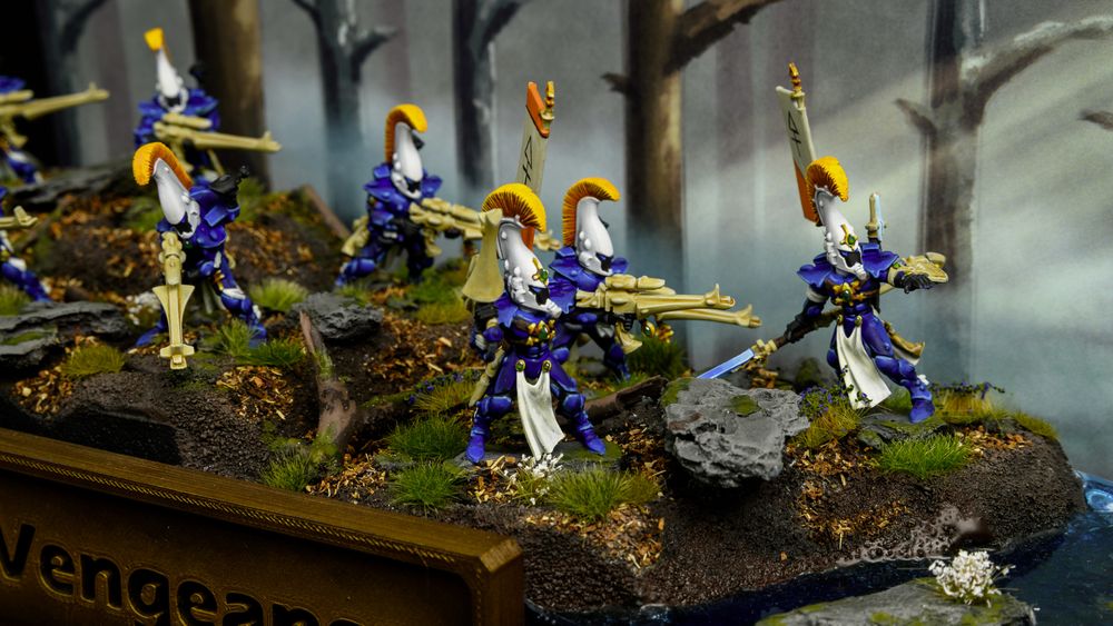 A picture of a miniatures display board featuring a woodland scene and painted backdrop with Dire Avenger miniatures. Grass, leaves and mossy rocks litter the forest floor, contrasting the Blue, white, and orange elf models.