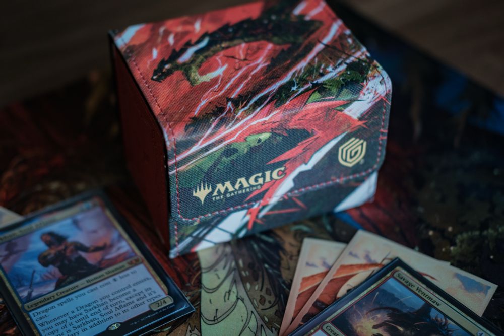 A Magic deckbox featuring a red and green illustration of a dragon. Spread around the deckbox are various Magic cards, the visible ones being dragon themed. They all sit on a gaming playmat.