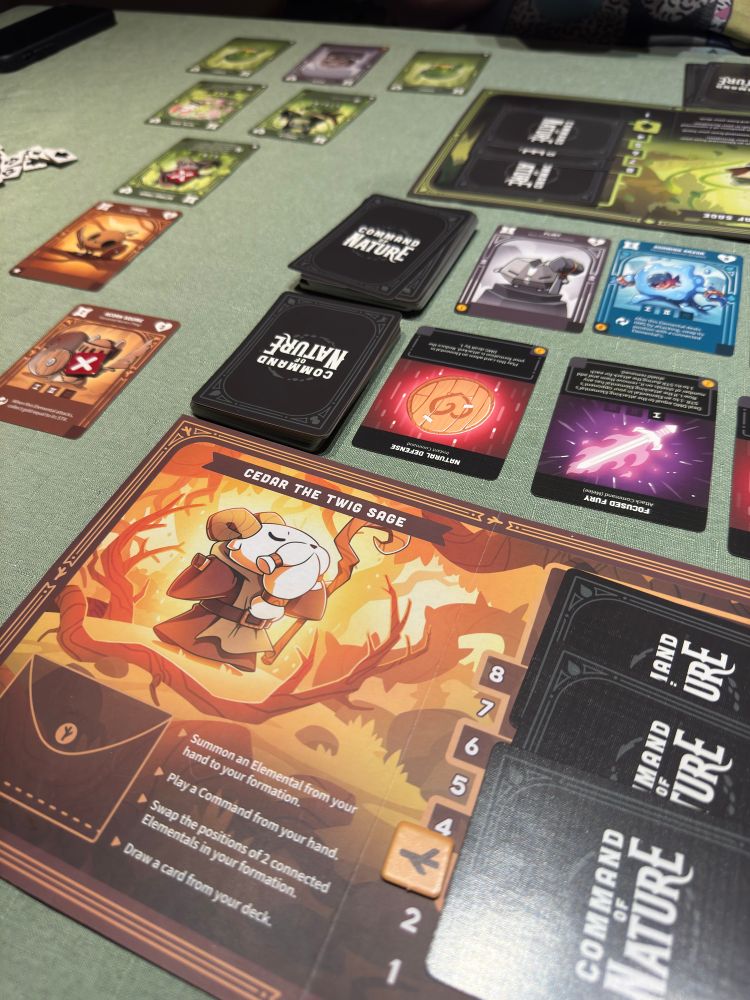 A picture showing off the game Command of Nature being played with a large game piece displaying a cartoonish illustration of a goat sage with various cards arranged around it.