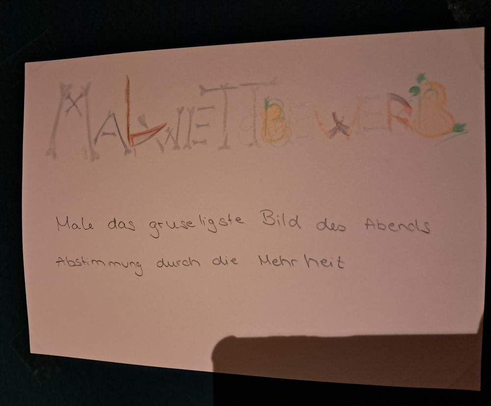 Foto von einem Papier mit dem handgeschriebenen Text "Malwettbewerb: Male das gruseligste Bild des Abends. Abstimmung durch die Mehrheit"