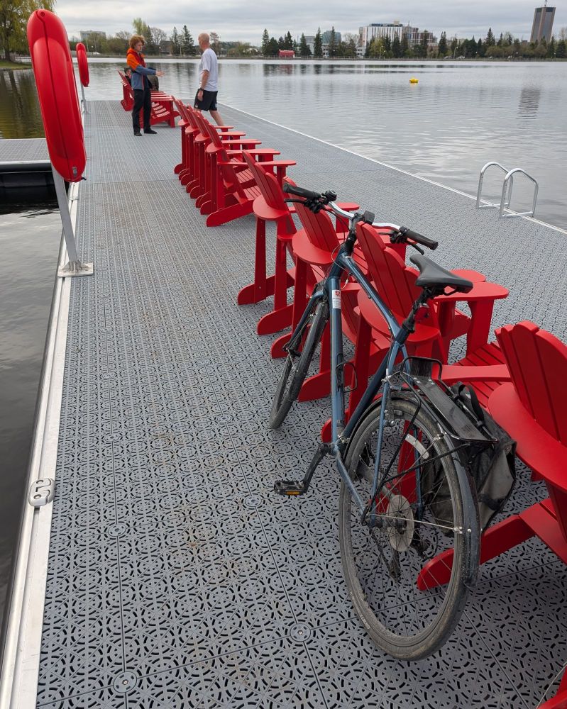New dock structure on Dow's Lake in Ottawa. The dock has a grey plastic deck, a long row of red Adirondack chairs, and swim ladders. In the background a Parks Canada employee talks to a passer-by. In the foreground a blue commuter bike leans against the chairs.